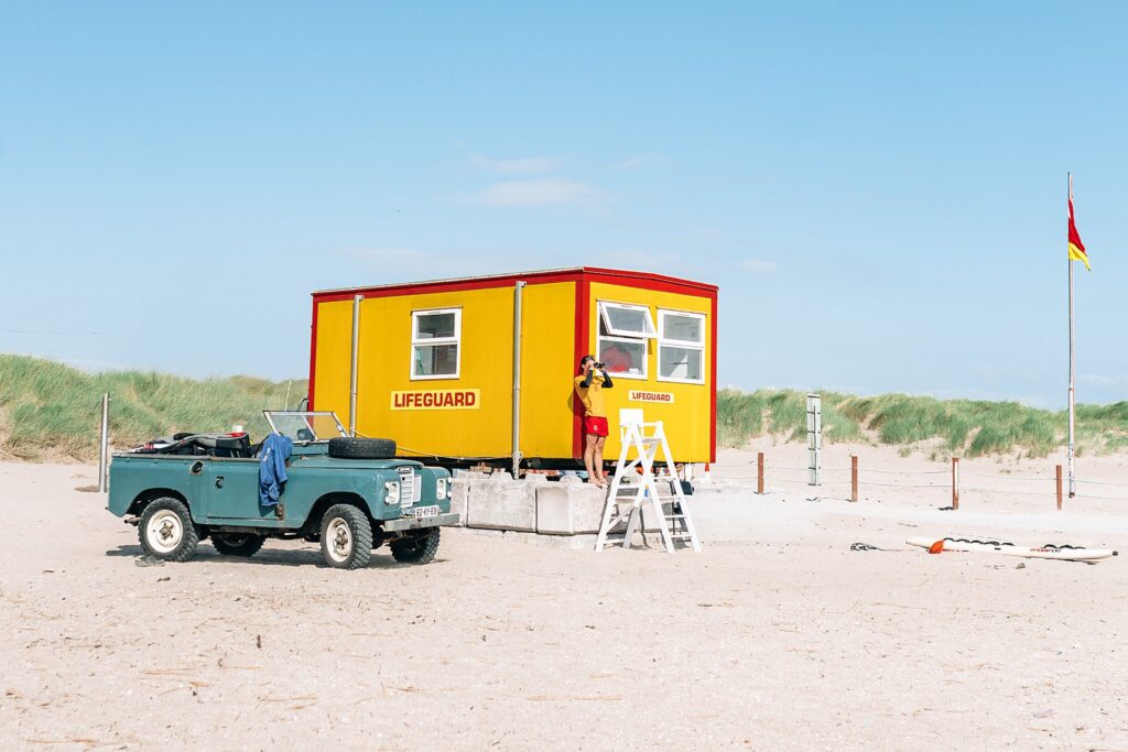 Classic life guard on duty in front of his yellow post and next to his muted blue convertible bronco on the beautiful clean, Inch beach with soft sand, muted grass and blue skies in the background.